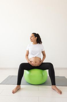 A young pregnant woman doing relaxation exercise using a fitness ball while sitting on a mat and holding her tummy