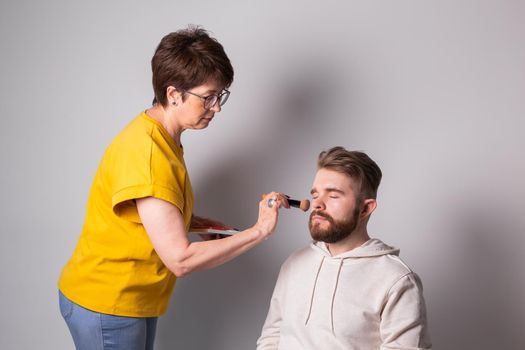 Bearded man getting makeup. Woman visagist works with brush