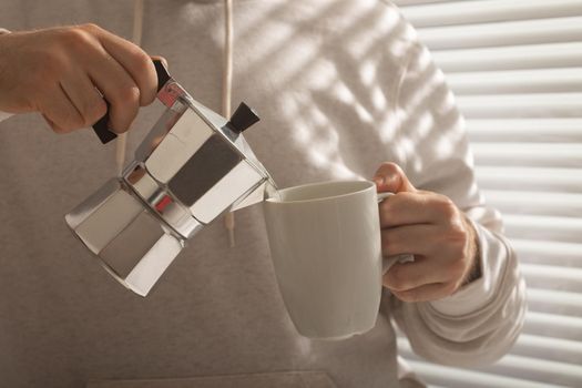 Close-up portrait of young stylish hipster male pouring coffee at office on summer day. Invigorating morning and positive mood