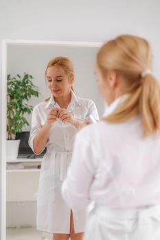 A blonde woman in white formal clothes looks at herself in the mirror. On a white background, green leaves at the back