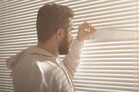 Rear view of young man with beard peeks through hole in the window blinds and looks out into the street. Surveillance and curiosity