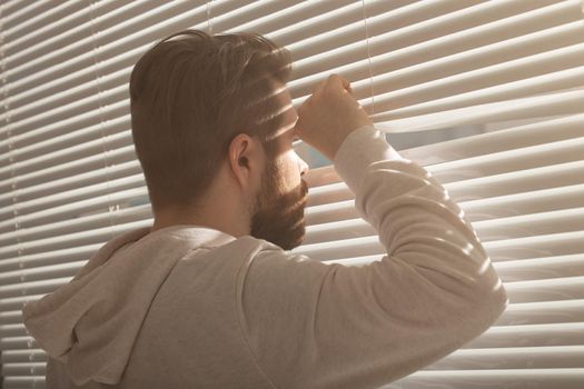 Rear view of young man with beard peeks through hole in the window blinds and looks out into the street. Surveillance and curiosity