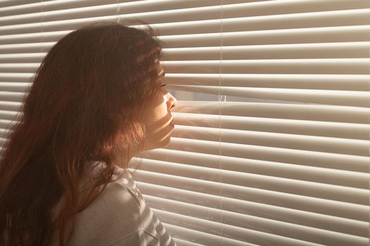 Rear view of beautiful young woman with long hair peeks through hole in the window blinds and looks out the window. Surveillance and curiosity