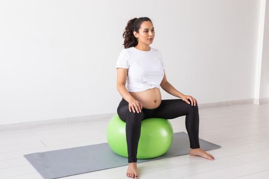 A young pregnant woman doing relaxation exercise using a fitness ball while sitting on a mat and holding her tummy