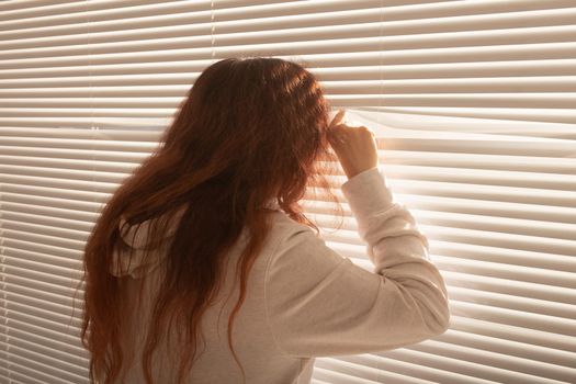 Rear view of beautiful young woman with long hair peeks through hole in the window blinds and looks out the window. Surveillance and curiosity