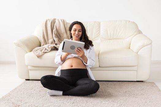 Pregnant woman holding tablet sitting on a carpet near a couch in the living room at home.