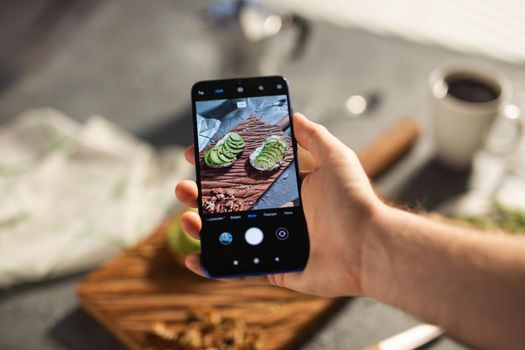 Hands take pictures on smartphone of two beautiful healthy sour cream and avocado sandwiches lying on board on the table. Social media and food