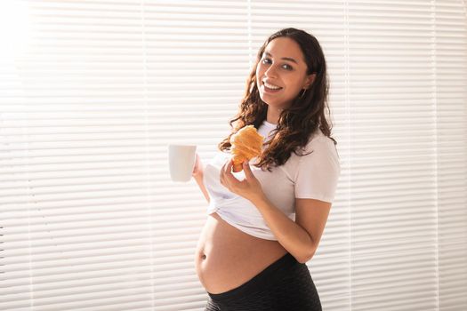 Beautiful pregnant woman holding croissant and cup of coffee in her hands during morning breakfast. Good health and positive attitude while expecting baby