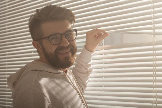 Rear view of young man with beard peeks through hole in the window blinds and looks out into the street. Surveillance and curiosity