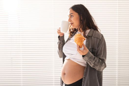Healthy beautiful pregnant woman drinking tea and eating croissant during lunch. High-calorie nutrition while waiting for birth of baby