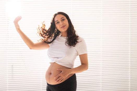 Smiling young beautiful pregnant woman touching her belly and hair and rejoicing. Health and thinking about the future while waiting for baby. Copyspace