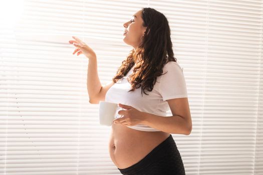 Curious young beautiful pregnant woman drinking tea and looking through the blinds at the window. Joy and good news while waiting for baby