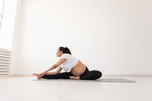 Young flexible pregnant woman doing gymnastics on rug on the floor on white background. Preparing the body for easy childbirth