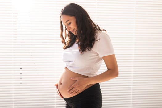 Smiling young beautiful pregnant woman touching her belly and rejoicing. Concept of positive and pleasant feelings while waiting for the baby.