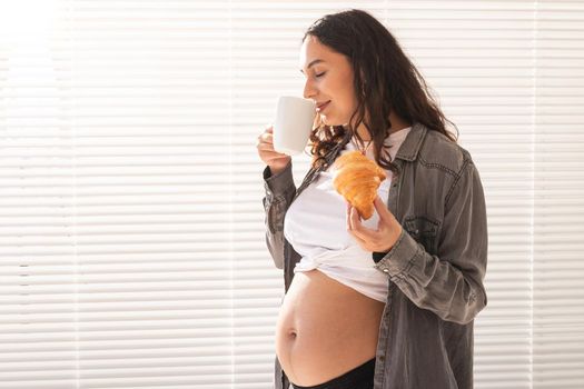 Healthy beautiful pregnant woman drinking tea and eating croissant during lunch. High-calorie nutrition while waiting for birth of baby