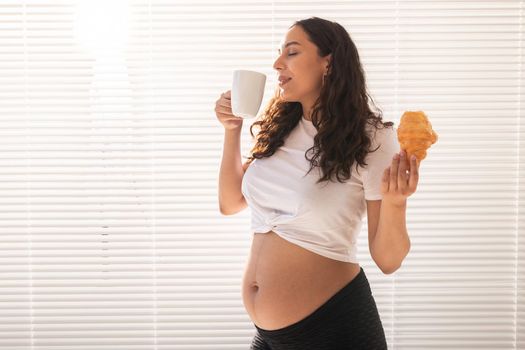 Pregnant woman eating croissant and drinks coffee. Pregnancy and maternity leave.