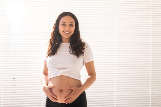 Smiling young beautiful pregnant woman touching her belly and rejoicing. Concept of positive and pleasant feelings while waiting for the baby.