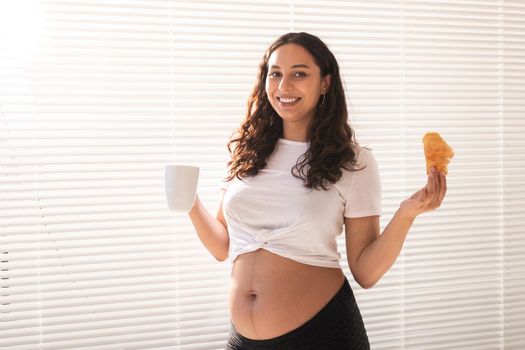 Beautiful pregnant woman holding croissant and cup of coffee in her hands during morning breakfast. Good health and positive attitude while expecting baby