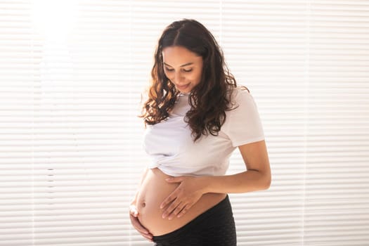 Smiling young beautiful pregnant woman touching her belly and rejoicing. Concept of positive and pleasant feelings while waiting for the baby.