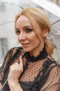 The blonde stands under a transparent umbrella during the rain. The fall season. Rear view. The woman is dressed in a black lace dress, her hair pulled back in a ponytail