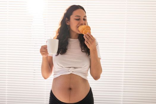 Pregnant woman eating croissant and drinks coffee. Pregnancy and maternity leave.