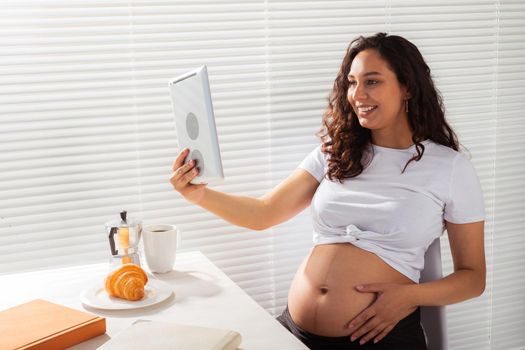 Happy pregnant young beautiful woman talking to mom using video call during morning breakfast. Communication and positive attitude during pregnancy