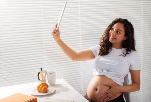 Happy pregnant young beautiful woman talking to mom using video call during morning breakfast. Communication and positive attitude during pregnancy