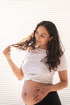 Smiling young beautiful pregnant woman touching her belly and hair and rejoicing. Health and thinking about the future while waiting for baby. Copyspace