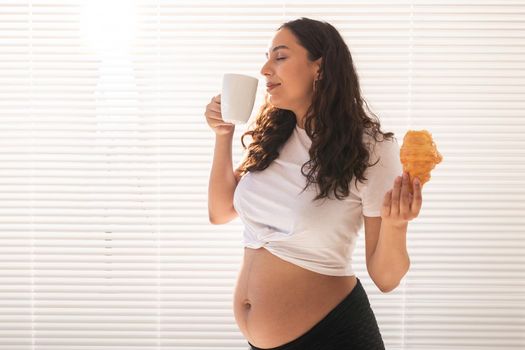 Beautiful pregnant woman holding croissant and cup of coffee in her hands during morning breakfast. Good health and positive attitude while expecting baby