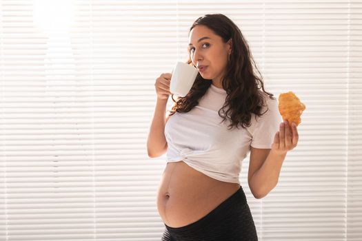 Pregnant woman eating croissant and drinks coffee. Pregnancy and maternity leave.