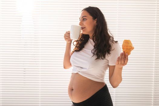 Beautiful pregnant woman holding croissant and cup of coffee in her hands during morning breakfast. Good health and positive attitude while expecting baby