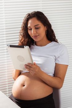 Hispanic pregnant woman using digital tablet while breakfast. Technology, pregnancy and maternity leave.