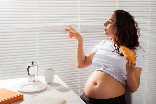Happy pregnant young beautiful woman eating croissant and looking througt blinds during morning breakfast. Pleasant morning and positive attitude during pregnancy