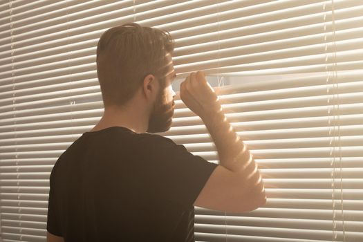 Rear view of young man with beard peeks through hole in the window blinds and looks out into the street. Surveillance and curiosity