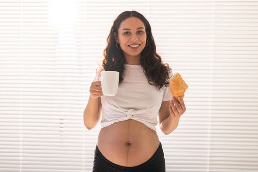 Pregnant woman eating croissant and drinks coffee. Pregnancy and maternity leave.