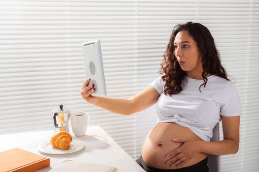 Happy pregnant young beautiful woman talking to mom using video call during morning breakfast. Communication and positive attitude during pregnancy