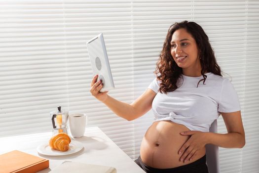 Happy pregnant young beautiful woman talking to mom using video call during morning breakfast. Communication and positive attitude during pregnancy