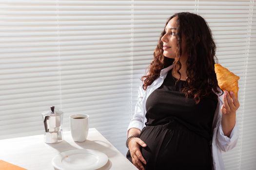 Joyful pregnant young beautiful woman looks through blinds during her morning breakfast with coffee and croissants. Good morning and waiting for meeting with a baby