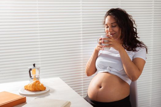 Joyful pregnant young beautiful woman looks through blinds during her morning breakfast with coffee and croissants. Good morning and waiting for meeting with a baby