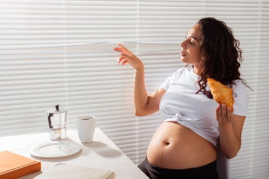 Happy pregnant young beautiful woman eating croissant and looking througt blinds during morning breakfast. Pleasant morning and positive attitude during pregnancy