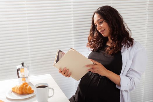 Positive young beautiful woman reads book during morning breakfast with coffee and croissants. Pleasant morning and expectation of meeting baby