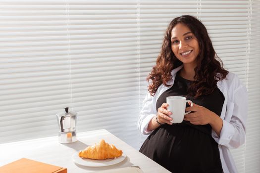 Joyful pregnant young beautiful woman looks through blinds during her morning breakfast with coffee and croissants. Good morning and waiting for meeting with a baby