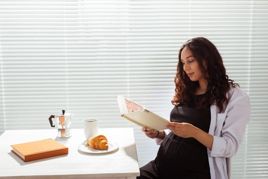 Side view of happy young beautiful woman reading book while having morning breakfast with coffee and croissants on background of blinds. Good morning and pleasant lunch break