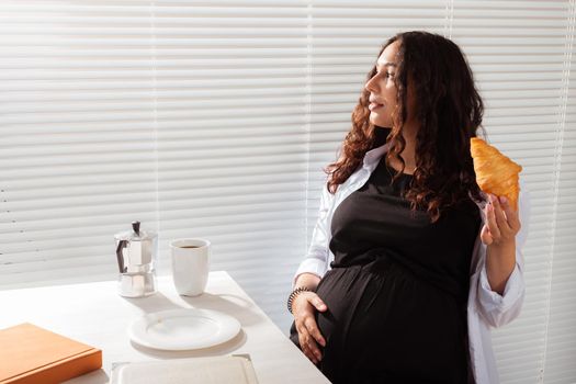 Happy pregnant young beautiful woman eating croissant and looking througt blinds during morning breakfast. Pleasant morning and positive attitude during pregnancy