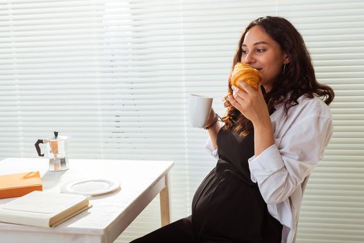 Happy pregnant young beautiful woman eating croissant and looking througt blinds during morning breakfast. Pleasant morning and positive attitude during pregnancy