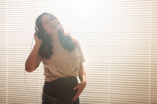 Curly-haired brunette pacified pregnant woman listens to pleasant classical music using smartphone and headphones. Concept of a soothing mood before meeting baby