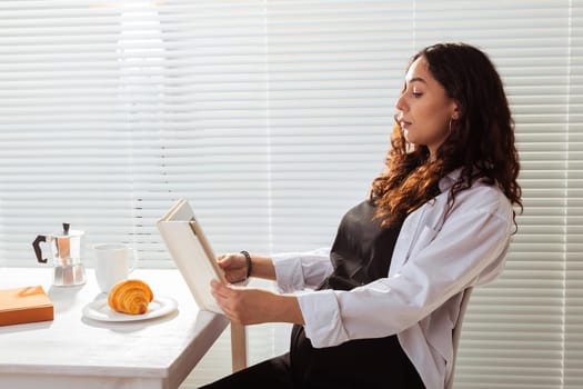 Side view of happy young beautiful woman reading book while having morning breakfast with coffee and croissants on background of blinds. Good morning and pleasant lunch break