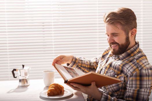 Side view of pensive young bearded hipster man reading book and having dinner with croissant and cup of coffee on background of blinds.