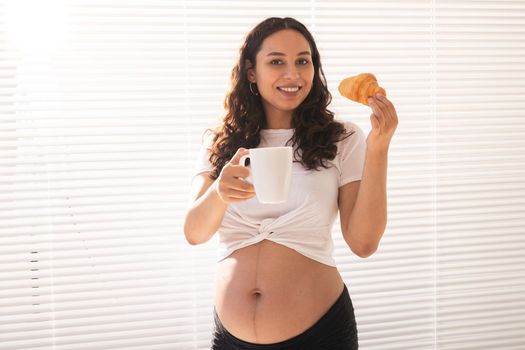 Beautiful pregnant woman holding croissant and cup of coffee in her hands during morning breakfast. Good health and positive attitude while expecting baby