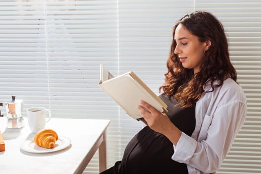 Side view of happy young beautiful woman reading book while having morning breakfast with coffee and croissants on background of blinds. Good morning and pleasant lunch break
